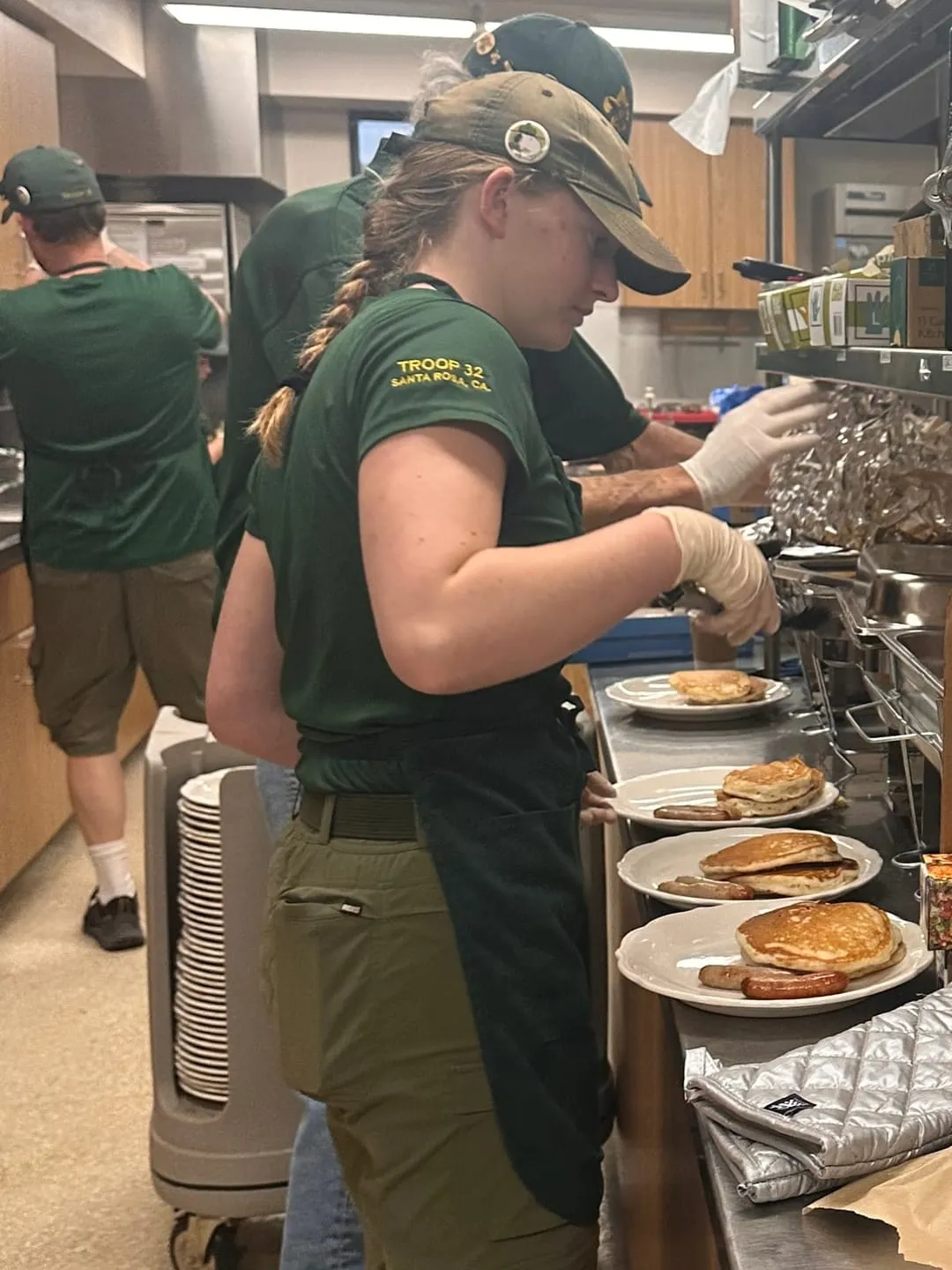 A Scout in a Troop 32 shirt, apron, and gloves plates pancakes and sausages on a busy kitchen serving line.