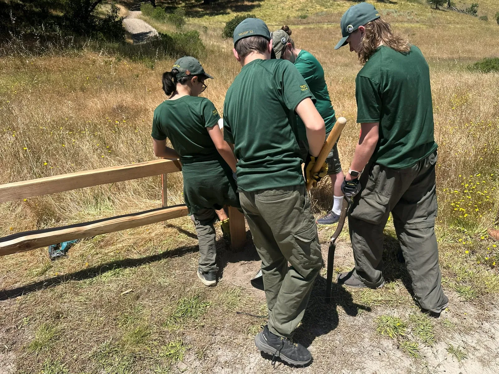 Scouts in Troop 32 caps set a wooden post for a split-rail fence beside a hillside trail.
