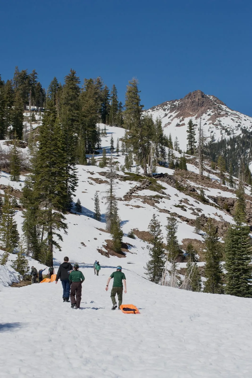 Scouts in green Troop 32 shirts haul orange sleds up a snowy slope toward a rocky mountain peak.
